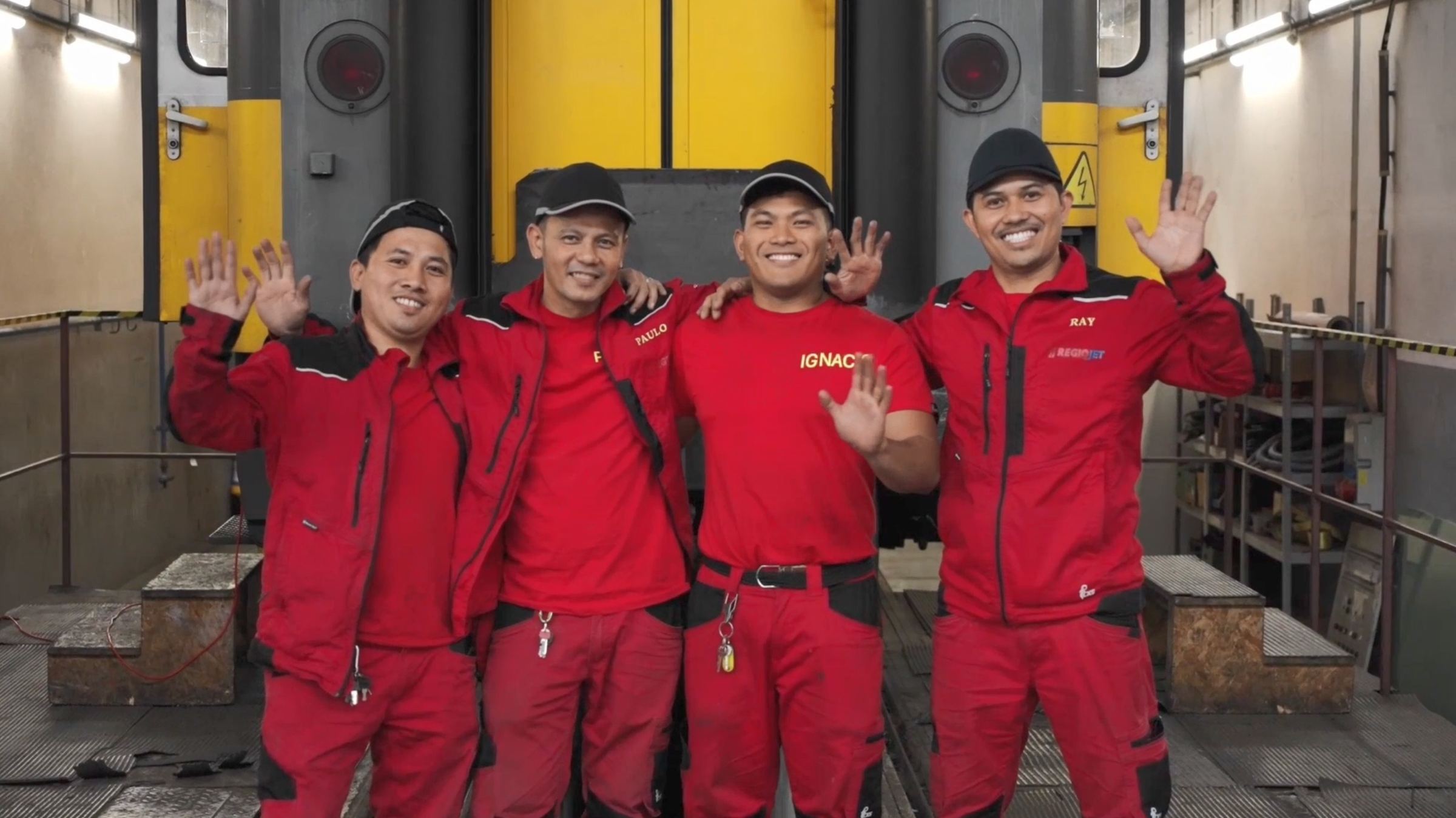 Four RegioJet maintenance workers in red workwear waving in the depot hall