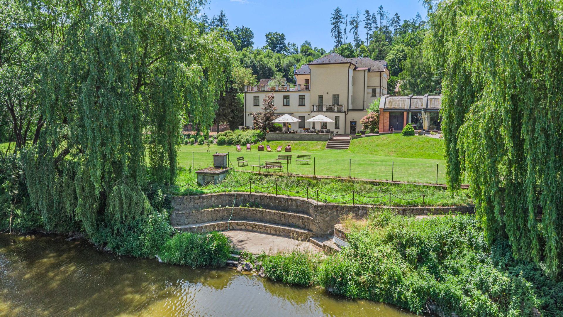 The accommodation building above the river, surrounded by willows and lawn